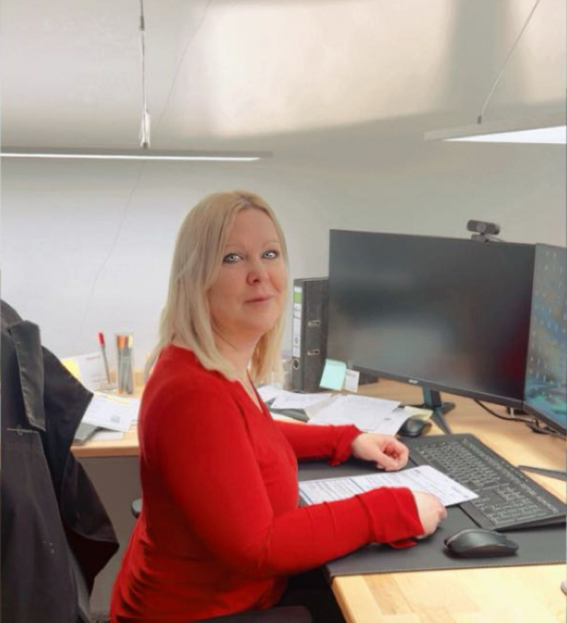 Büromitarbeiterin Aylin Kay sitzt an ihrem Schreibtisch mit Computer und schaut in die Kamera.