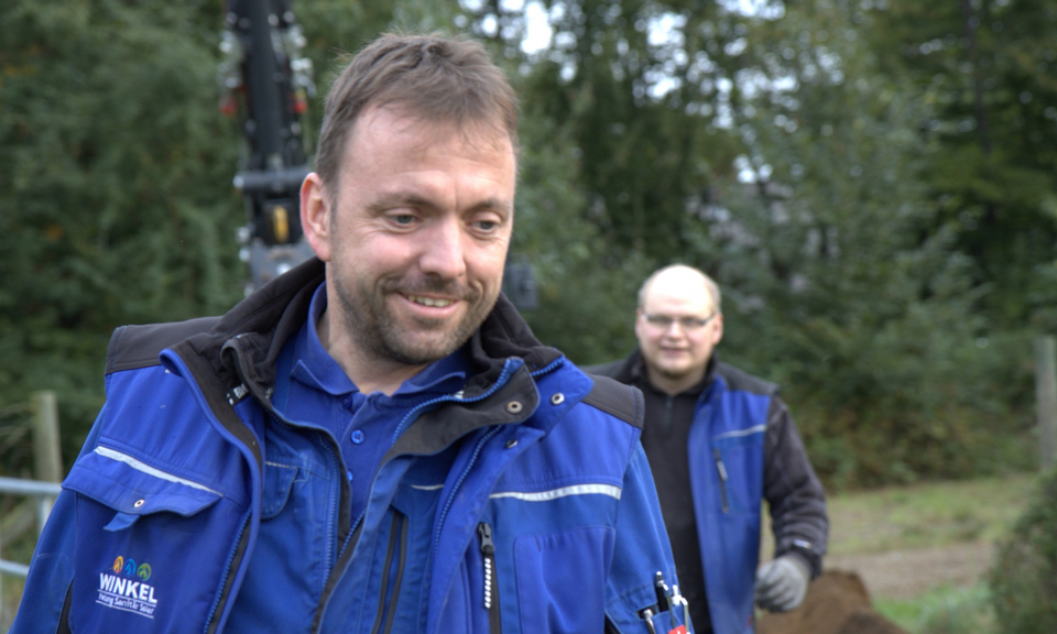 Thomas Winkel, Inhaber der Winkel GmbH, lächelt im Vordergrund in blauer Firmenjacke, im Hintergrund ein weiterer Mitarbeiter auf einer Baustelle im Freien.