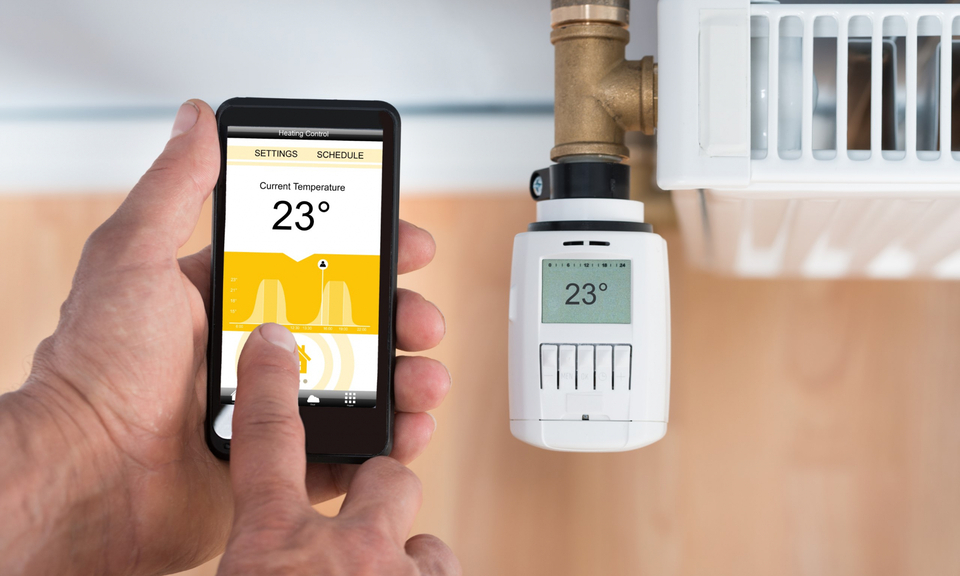 Hand hält ein Smartphone mit Heizungssteuerungs-App neben einem Heizkörperthermostat.